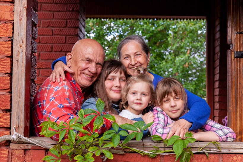 Abuelos sonriendo junto a sus tres nietas asomados al marco de una ventana de ladrillo con plantas verdes.