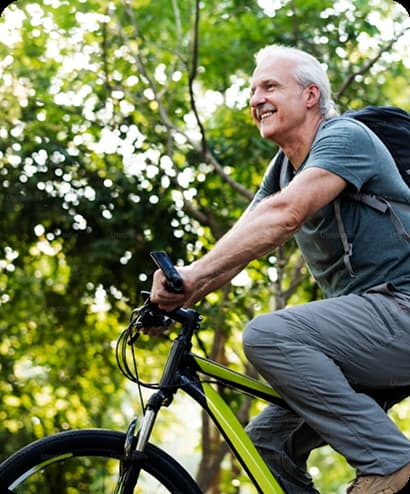 Foto de adulto mayor sonriendo mientras monta en bicicleta de montaña al aire libre rodeado de árboles y naturaleza.