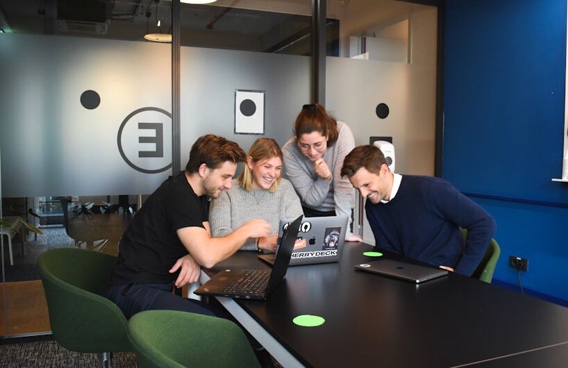 Cuatro compañeros de trabajo sonriendo y colaborando juntos alrededor de una computadora portátil en una sala de reuniones de oficina moderna.