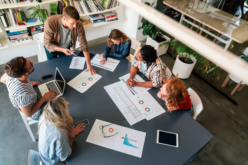 Vista superior de un equipo de trabajo en reunión de negocios alrededor de una mesa compartida, analizando gráficos impresos, datos y reportes en una oficina moderna.