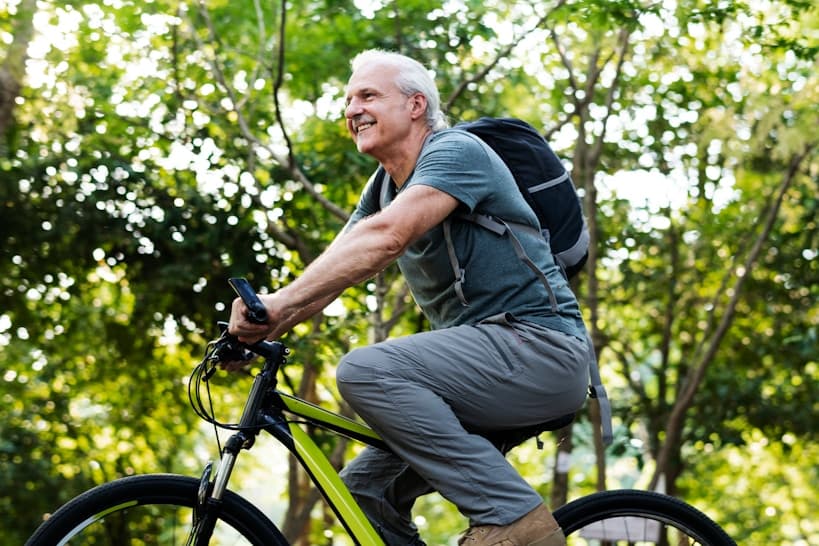 Hombre adulto mayor sonriendo mientras anda en bicicleta por un sendero al aire libre rodeado de árboles, llevando una mochila en la espalda.