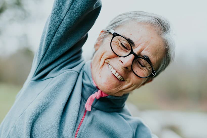 Foto de mujer adulta mayor con lentes y cabello canoso sonriendo con los ojos cerrados mientras estira un brazo hacia arriba, vistiendo un abrigo polar celeste al aire libre.