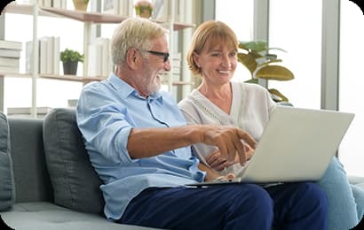 Pareja de adultos mayores sonríe mientras utiliza una computadora portátil sentada en un sofá.